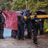 RCMP officers hold a tarp to block the view of an arrest from reporters and the public while one officer directs a photographer to move back to a position where they cannot see beyond the tarp. 